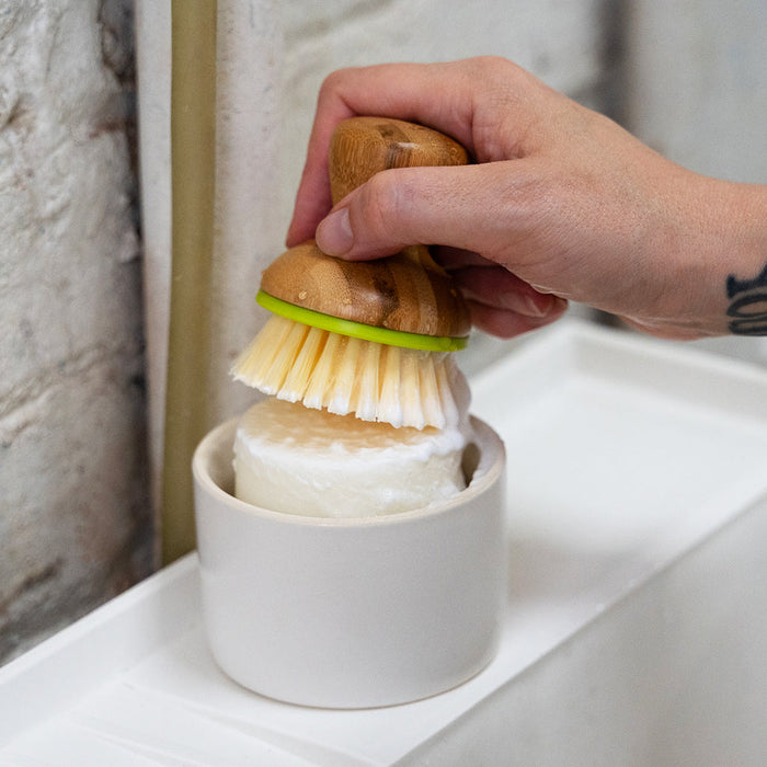 A photo of a person scrubbing a wet dish soap bar with a dish brush to create lather.